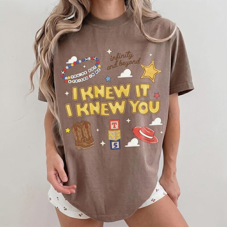 May include: A brown short-sleeve t-shirt featuring a graphic print with the text "I KNEW IT I KNEW YOU", "infinity and beyond", and "YOU'VE GOT A FRIEND IN ME". The design includes illustrations of a beaded bracelet, a star, cowboy boots, blocks, and a hat.