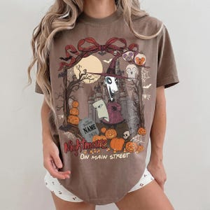 May include: A brown t-shirt featuring a graphic print of a tall, thin character in a purple hat, a tombstone with the text NAME 2026, pumpkins, a castle, and bats. The text Nightmare ON MAIN STREET is printed in red and white below the illustration.