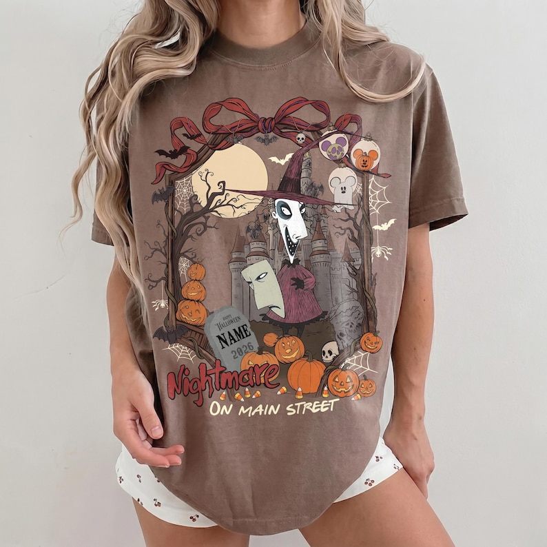 May include: A brown t-shirt featuring a graphic print of a tall, thin character in a purple hat, a tombstone with the text NAME 2026, pumpkins, a castle, and bats. The text Nightmare ON MAIN STREET is printed in red and white below the illustration.