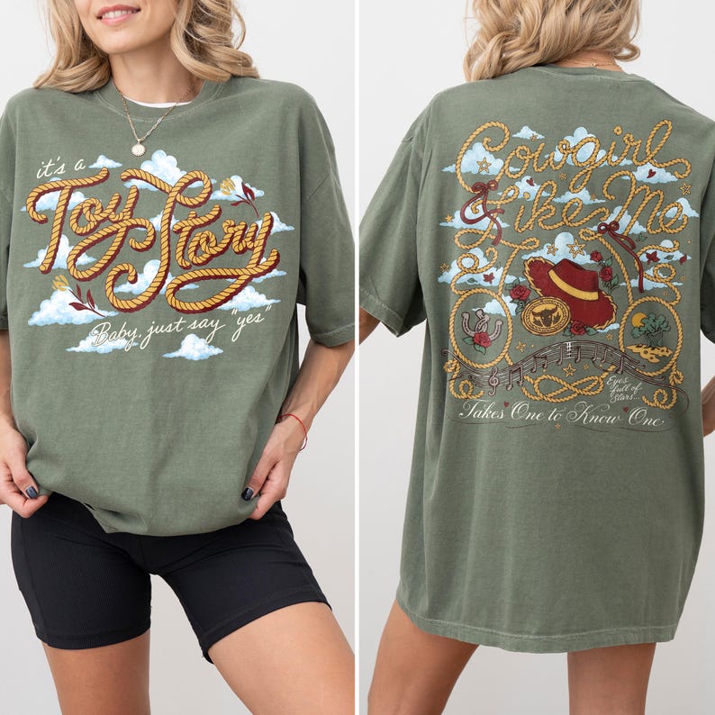 Op de afbeelding: Een olijfgroen t-shirt met een afbeelding op de voorkant met touwachtige tekst "it's a Toy Story Baby, just say "yes"" boven wolken. De achterkant toont een touwontwerp, een rode hoed, een hoefijzer en de tekst "Cowgirl like Me".