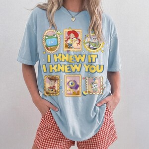 May include: A light blue t-shirt featuring a graphic print of Toy Story characters in postage stamp frames. The text reads I KNEW IT I KNEW YOU in yellow block letters. The design includes images of Jessie, Buzz Lightyear, Woody, and other characters.