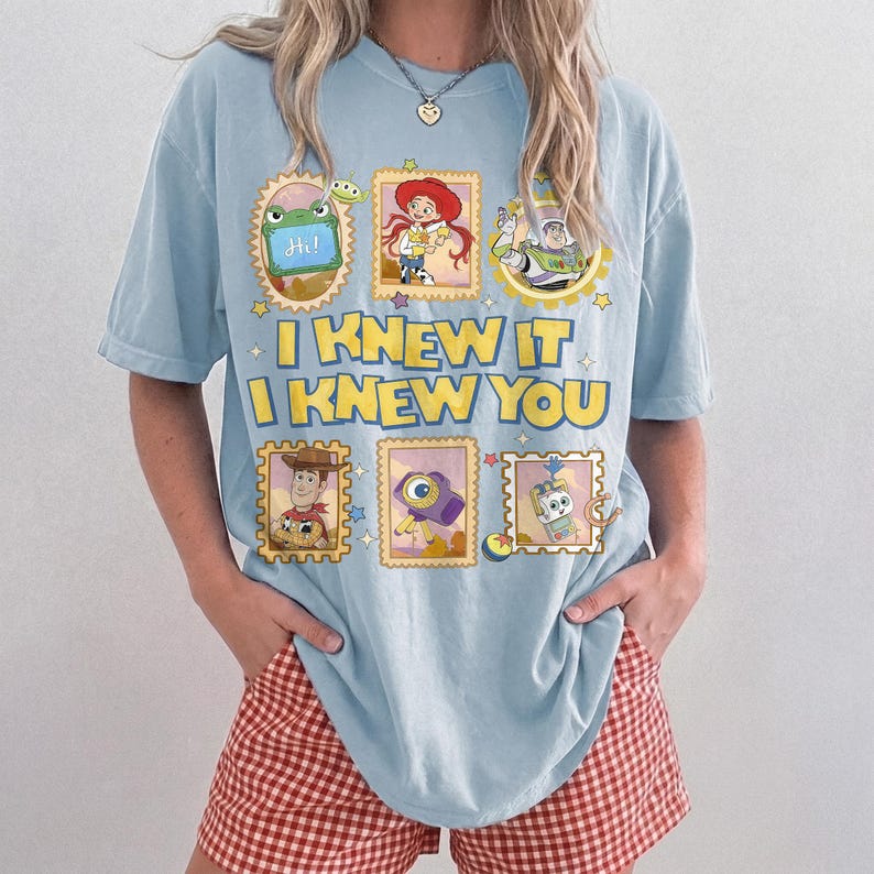 May include: A light blue t-shirt featuring a graphic print of Toy Story characters in postage stamp frames. The text reads I KNEW IT I KNEW YOU in yellow block letters. The design includes images of Jessie, Buzz Lightyear, Woody, and other characters.