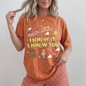 May include: An orange short-sleeve t-shirt featuring a graphic print with the text "I KNEW IT I KNEW YOU", "infinity and beyond", and "YOU'VE GOT A FRIEND IN ME". The design includes illustrations of a cowboy boot, a red hat, and toy blocks.