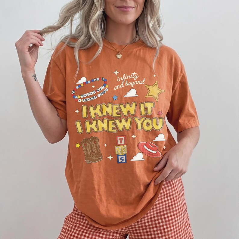 May include: An orange short-sleeve t-shirt featuring a graphic print with the text "I KNEW IT I KNEW YOU", "infinity and beyond", and "YOU'VE GOT A FRIEND IN ME". The design includes illustrations of a cowboy boot, a red hat, and toy blocks.