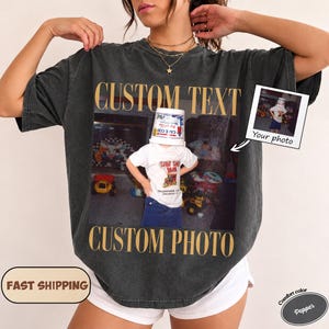 Può includere: Una t-shirt a maniche corte grigio antracite con una grande stampa grafica rettangolare. La stampa mostra la fotografia di un bambino con un secchio in testa, circondato da giocattoli. Il testo CUSTOM TEXT e CUSTOM PHOTO è in oro.
