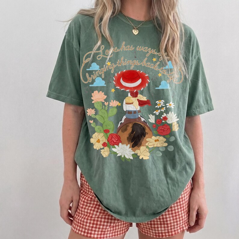 May include: A sage green short-sleeve t-shirt featuring a graphic of a cowgirl figure on a horse, surrounded by cacti and flowers. The text reads, "Hope has ways of bringing things back to you." The design includes a red hat and blue clouds.