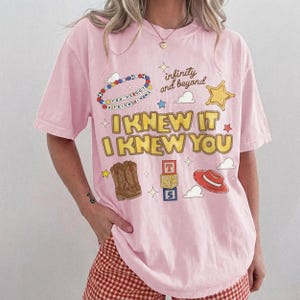 May include: A light pink short-sleeve t-shirt featuring a graphic print with the text "I KNEW IT I KNEW YOU", "infinity and beyond", and "YOU'VE GOT A FRIEND IN ME". The design includes illustrations of cowboy boots, a red hat, and blocks.