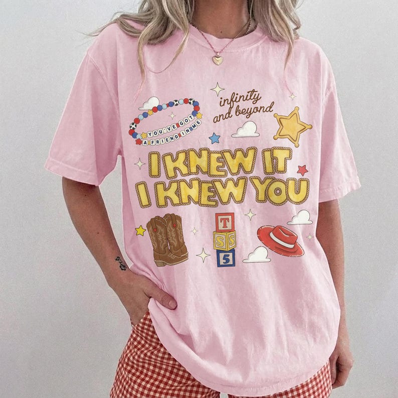 May include: A light pink short-sleeve t-shirt featuring a graphic print with the text "I KNEW IT I KNEW YOU", "infinity and beyond", and "YOU'VE GOT A FRIEND IN ME". The design includes illustrations of cowboy boots, a red hat, and blocks.