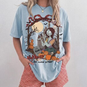May include: A light blue t-shirt featuring a graphic of a female character with long brown hair, a castle, a full moon, and pumpkins. The text reads "NAME 2026" on a tombstone and "Nightmare ON MAIN STREET" at the bottom, framed by a red bow.