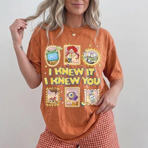 May include: An orange short-sleeve t-shirt featuring a graphic print of various Toy Story characters in postage stamp-style frames. The text "I KNEW IT I KNEW YOU" is printed in large yellow letters across the center of the shirt design.