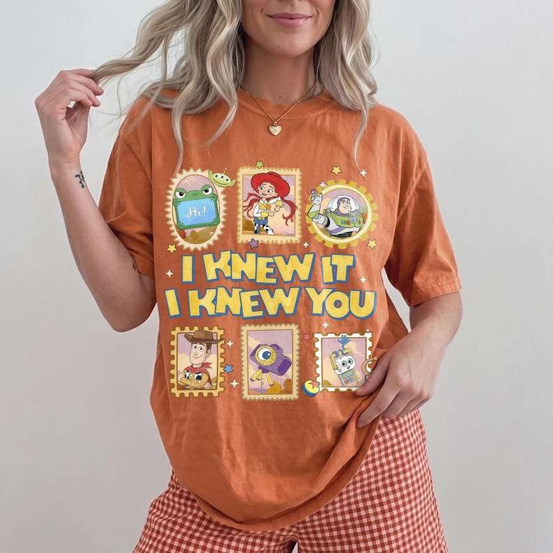 May include: An orange short-sleeve t-shirt featuring a graphic print of various Toy Story characters in postage stamp-style frames. The text "I KNEW IT I KNEW YOU" is printed in large yellow letters across the center of the shirt design.