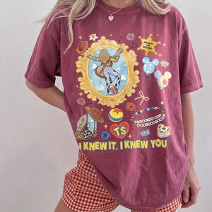 May include: A maroon short-sleeve t-shirt featuring a graphic print with a gold ornate frame, a person with a guitar, a sheriff badge with the text INFINITY AND BEYOND, Mickey-shaped balloons, a cowboy boot, a ball, and YOU'VE GOT A FRIEND IN ME.