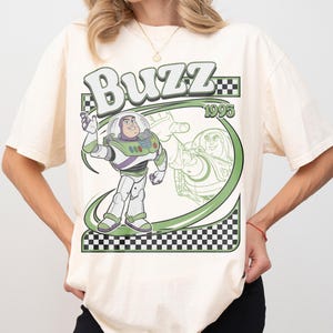 May include: Cream-colored t-shirt featuring a graphic of Buzz Lightyear from Toy Story. The design includes the word "Buzz" in a stylized font, the year "1995", and a green and black checkered border. Buzz Lightyear is depicted in his space suit.