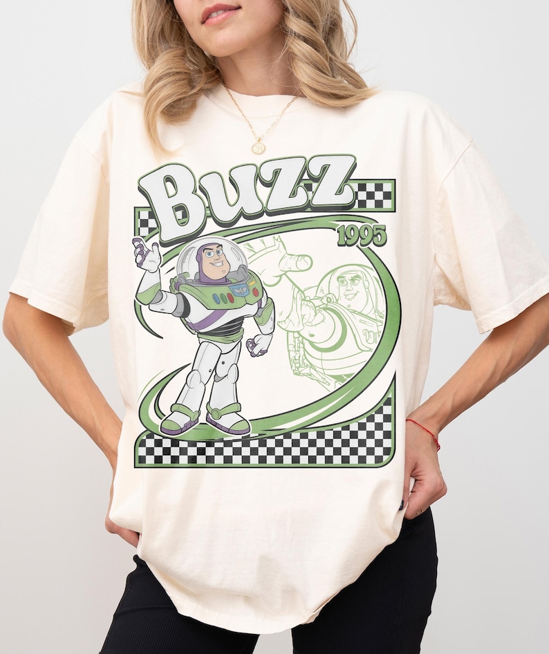 May include: Cream-colored t-shirt featuring a graphic of Buzz Lightyear from Toy Story. The design includes the word "Buzz" in a stylized font, the year "1995", and a green and black checkered border. Buzz Lightyear is depicted in his space suit.