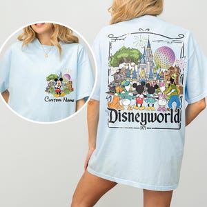 May include: Light blue t-shirt featuring a colorful Disneyworld graphic on the back and a smaller Mickey Mouse design on the front. The back graphic includes the text "Disneyworld" and "1971".