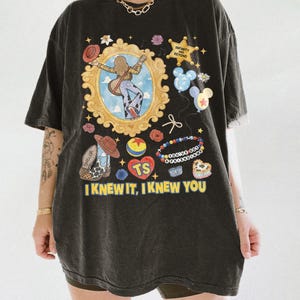 May include: A black oversized t-shirt featuring a graphic print with a gold ornate frame containing a figure with a guitar, a sheriff badge reading INFINITY AND BEYOND, balloons, a beaded bracelet, and the text I KNEW IT, I KNEW YOU.