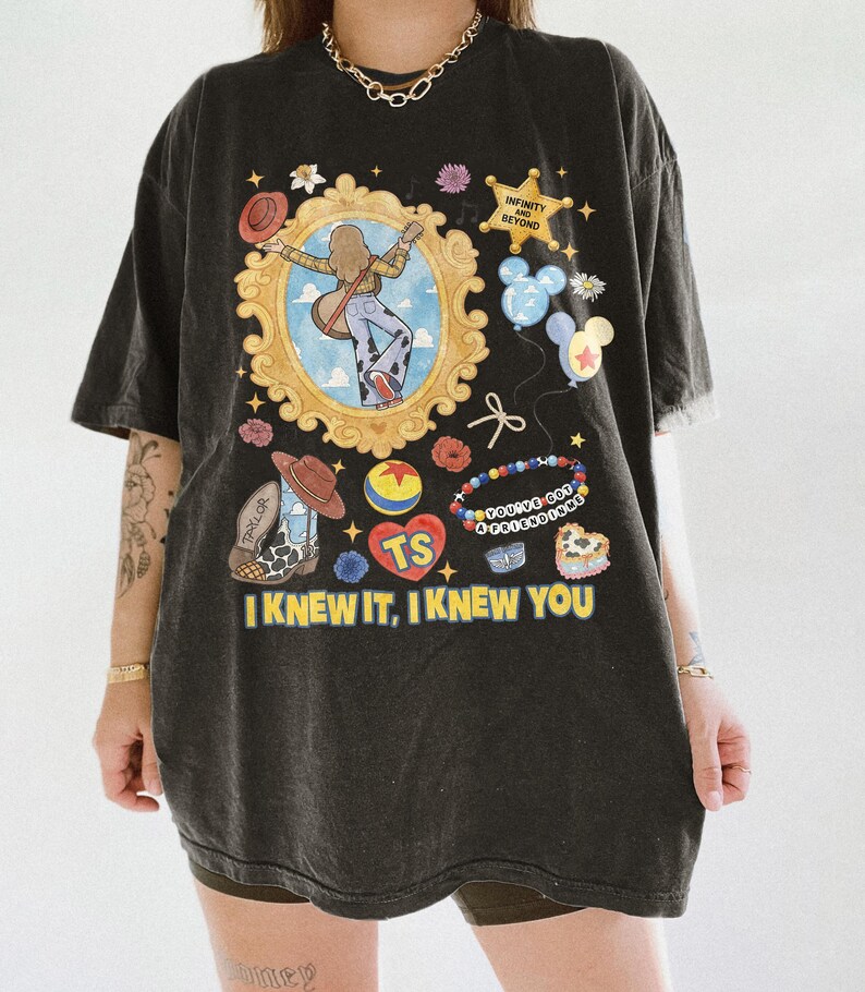May include: A black oversized t-shirt featuring a graphic print with a gold ornate frame containing a figure with a guitar, a sheriff badge reading INFINITY AND BEYOND, balloons, a beaded bracelet, and the text I KNEW IT, I KNEW YOU.