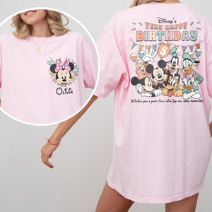 Puede incluir: Camiseta rosa claro con Minnie Mouse y el nombre "Olivia" en la parte delantera. La parte trasera de la camiseta muestra personajes de Disney con el texto "Disney's Very Happy Birthday" y "Wishing you a year filled with joy and good memories."