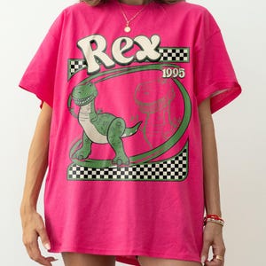 May include: A hot pink t-shirt featuring a green dinosaur graphic with the name "Rex" in white and a checkered pattern. The year "1995" is also displayed. The shirt is a casual, oversized fit.