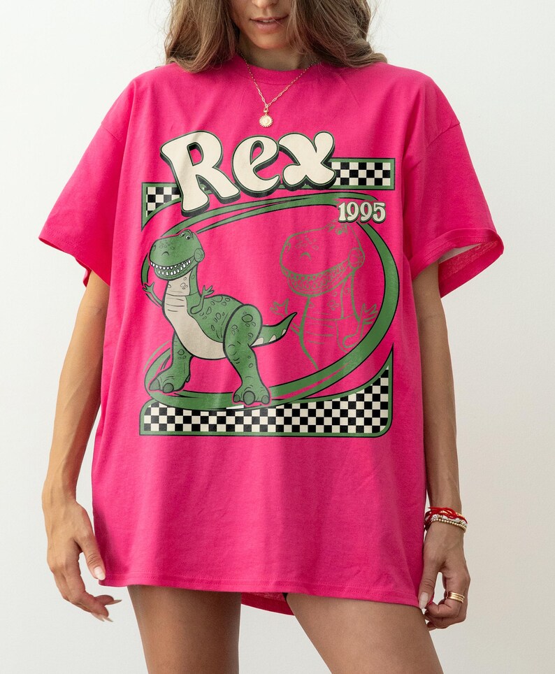 May include: A hot pink t-shirt featuring a green dinosaur graphic with the name "Rex" in white and a checkered pattern. The year "1995" is also displayed. The shirt is a casual, oversized fit.