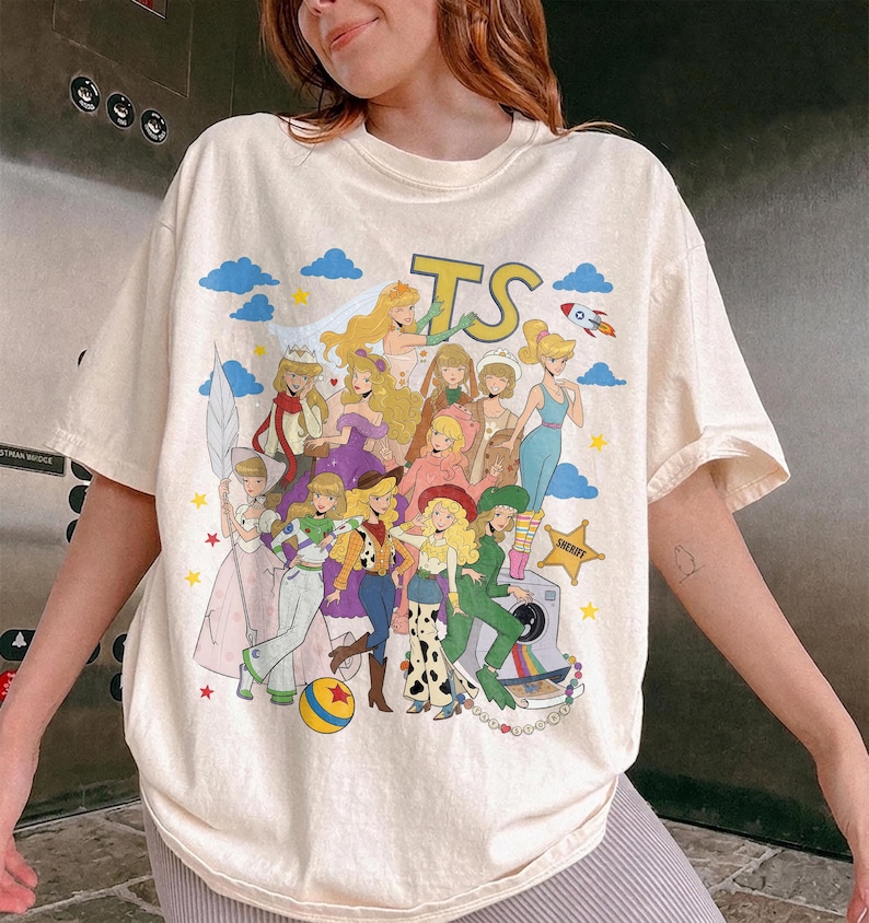 May include: A cream-colored short-sleeve t-shirt featuring a large graphic print of various animated characters dressed in costumes. The design includes the letters TS, a rocket, a yellow ball, and a sheriff star, all set against a background of blue clouds.