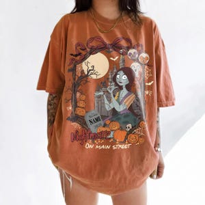 May include: An orange short-sleeve t-shirt featuring a graphic print of a female character with long dark hair, a castle, pumpkins, bats, and a tombstone with the text NAME 2026. The design includes a large bow at the top and the text Nightmare ON MAIN STREET.