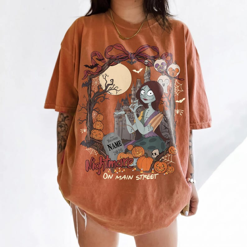 May include: An orange short-sleeve t-shirt featuring a graphic print of a female character with long dark hair, a castle, pumpkins, bats, and a tombstone with the text NAME 2026. The design includes a large bow at the top and the text Nightmare ON MAIN STREET.