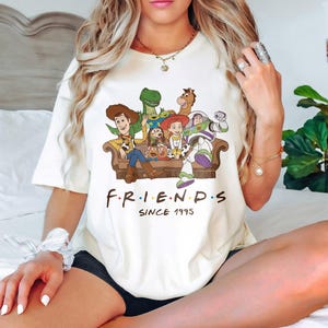May include: Cream-colored t-shirt featuring a graphic of Toy Story characters on a brown couch. The word "FRIENDS" is printed below the graphic, with "SINCE 1995" underneath. The shirt has short sleeves and a crew neck.