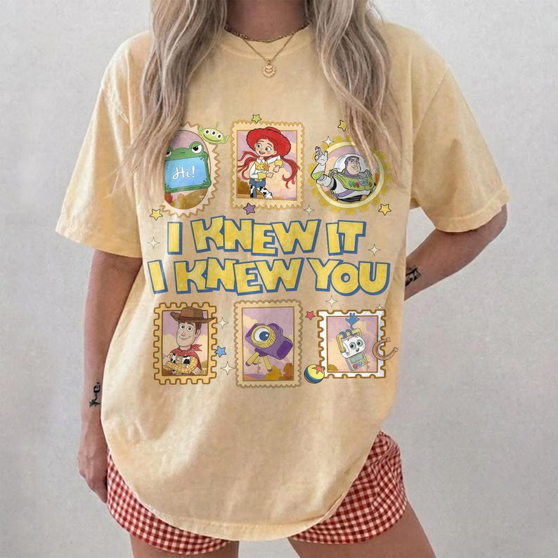 May include: A pale yellow short-sleeve t-shirt featuring a graphic print of Toy Story characters in postage stamp frames. The text reads I KNEW IT I KNEW YOU in bold blue letters. The design includes illustrations of Rex, Jessie, Buzz Lightyear, Woody, a camera, and Forky.