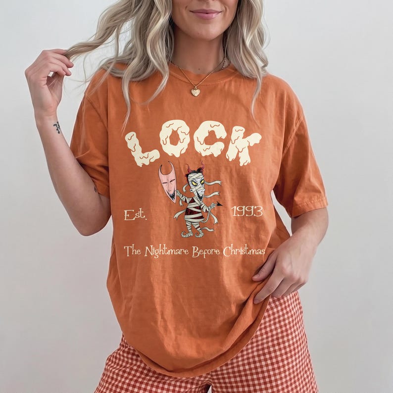 Op de afbeelding: Oranje T-shirt met het woord "LOCK" in een speels lettertype, en een mummie-achtig personage met een schild. De tekst "Est. 1993" en "The Nightmare Before Christmas" staan ook op het shirt.