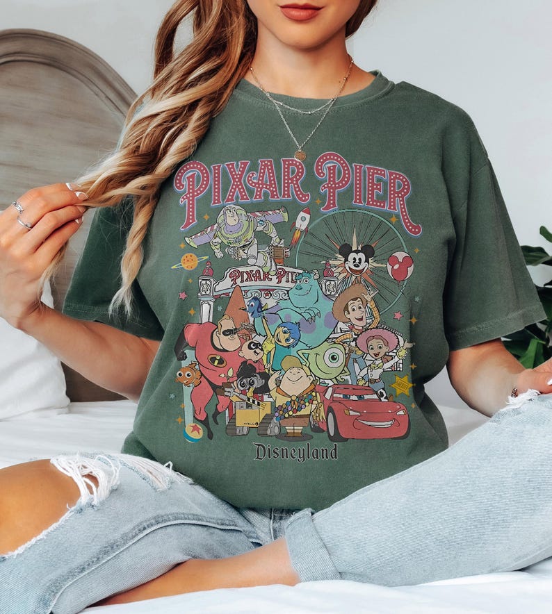 May include: Green t-shirt with a vibrant graphic of Pixar characters, such as Buzz Lightyear and Mike Wazowski. The design includes the words "Pixar Pier" and "Disneyland" in a retro style.