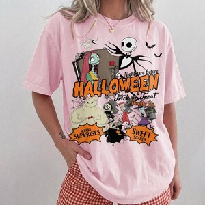 K&ouml;nnte beinhalten: Ein hellrosa T-Shirt mit einem Grafikdruck der Charaktere aus Nightmare Before Christmas. Das Design zeigt Jack Skellington, Sally und Oogie Boogie mit dem Text Nightmare Before Halloween, Trick or Treat Candy Co., Sugrry Surprises und Sweet Scares in orangefarbener und schwarzer Schrift.