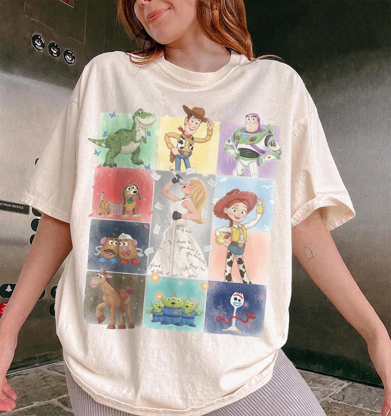 K&ouml;nnte beinhalten: Ein cremefarbenes T-Shirt mit kurzen &Auml;rmeln und einem Raster aus Toy Story-Figuren, darunter Rex, Woody, Buzz Lightyear, Slinky Dog, Mr. und Mrs. Potato Head, Bullseye, die Aliens, Jessie, Forky sowie eine mittige Illustration einer Frau.