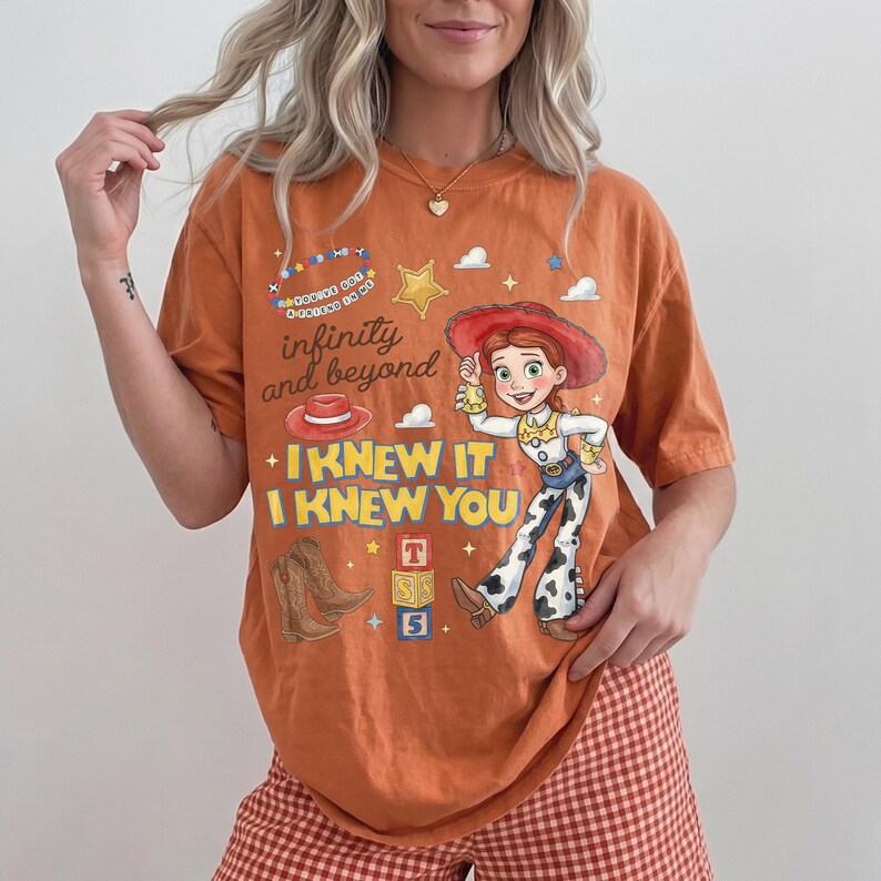 Pode incluir: Uma t-shirt laranja com uma estampa gr&aacute;fica de uma figura de cowgirl, botas de cowboy castanhas, um chap&eacute;u vermelho e blocos de alfabeto. O texto diz "infinity and beyond" e "I KNEW IT I KNEW YOU" com pequenas estrelas e nuvens.