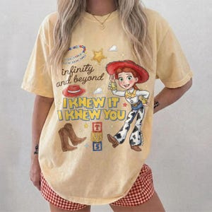 Pode incluir: Uma t-shirt de manga curta amarelo-p&aacute;lido com um gr&aacute;fico de uma cowgirl, um chap&eacute;u de cowboy vermelho, botas castanhas e blocos de letras. O texto diz "infinity and beyond", "I KNEW IT" e "I KNEW YOU" junto a estrelas e pequenas nuvens.