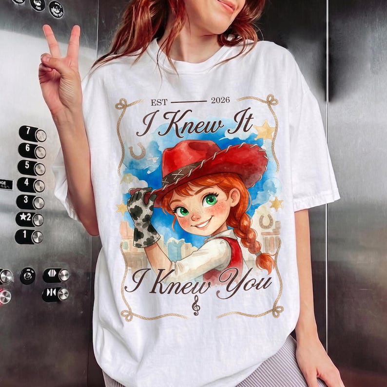 May include: A white t-shirt featuring a graphic of a cartoon character with red hair wearing a red cowboy hat and a cow-print glove. The text reads EST 2026, I Knew It, and I Knew You, framed by a rope design against a blue watercolor background.