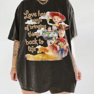 Pu&ograve; includere: Una t-shirt nera a maniche corte con una stampa grafica di Woody, Buzz Lightyear e Jessie di Toy Story. Il testo recita "Love has ways of bringing things back to life", accompagnato da un'illustrazione in stile acquerello di un albero e colline.