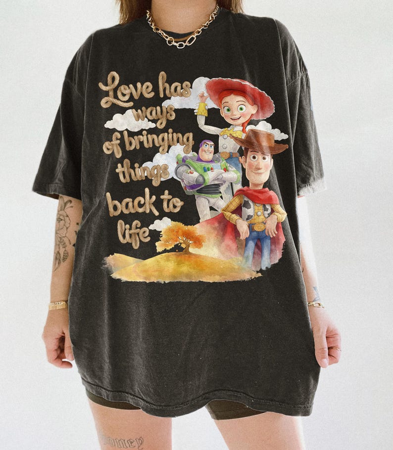 Pu&ograve; includere: Una t-shirt nera a maniche corte con una stampa grafica di Woody, Buzz Lightyear e Jessie di Toy Story. Il testo recita "Love has ways of bringing things back to life", accompagnato da un'illustrazione in stile acquerello di un albero e colline.