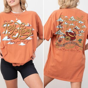Op de afbeelding: Een oranje t-shirt met korte mouwen voorzien van een voorzijde met touwachtige tekst it's a Toy Story Baby, just say yes en een achterzijde met de tekst Cowgirl like Me Takes One to Know One, inclusief een hoed en een lasso.