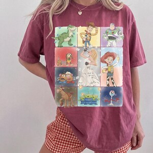 K&ouml;nnte beinhalten: Ein dunkelrotes Kurzarm-T-Shirt mit einem Raster aus Toy Story-Figuren, darunter Rex, Woody, Buzz Lightyear, Slinky Dog, Mr. und Mrs. Potato Head, Bullseye, die Aliens und Forky, um eine zentrale Illustration einer S&auml;ngerin in einem Kleid.