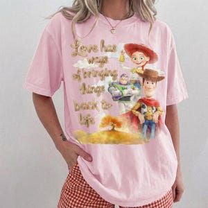 Pu&ograve; includere: Una t-shirt rosa chiaro con una stampa grafica di Woody, Buzz Lightyear e Jessie di Toy Story. Il testo "Love has ways of bringing things back to life" &egrave; posizionato sopra un'illustrazione paesaggistica con un albero e delle nuvole.