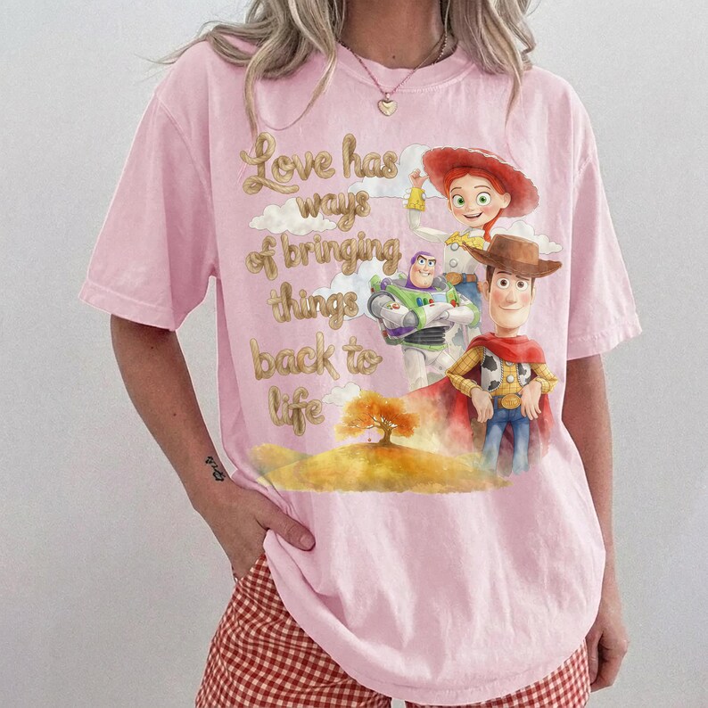 Pu&ograve; includere: Una t-shirt rosa chiaro con una stampa grafica di Woody, Buzz Lightyear e Jessie di Toy Story. Il testo "Love has ways of bringing things back to life" &egrave; posizionato sopra un'illustrazione paesaggistica con un albero e delle nuvole.