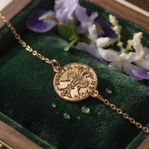Könnte beinhalten: Ein goldfarbenes Armband mit einem runden Anhänger, der ein geprägtes Fleur-de-Lis-Motiv zeigt. Die feine Kette ist an beiden Seiten des münzförmigen Anhängers befestigt, der detaillierte Texturen und Schriftzüge am äußeren Rand aufweist.