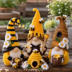 May include: Three crocheted gnomes and a small beehive. The left gnome has a yellow and black striped hat with white daisies. The center gnome wears a yellow hat with a honey drip design and holds a honey dipper. The right gnome has a brown hat with daisies and holds a honey pot. A small yellow and black beehive sits in front.