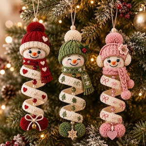 May include: Three hanging snowman ornaments with crocheted bodies, each wearing a knit hat and scarf. The left snowman has red accents and hearts, the center has green accents and trees, and the right has pink accents and bows, all with spiral tails.