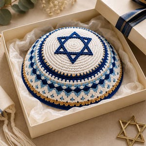 May include: A white and blue crocheted kippah featuring a prominent Star of David at the center. The dome-shaped head covering displays intricate geometric patterns in shades of navy blue, light blue, and gold, presented inside a gift box.