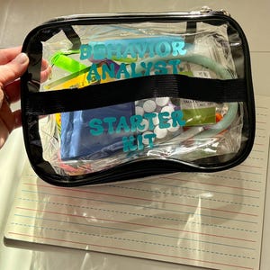 Puede incluir: Una bolsa transparente con cremallera con la inscripción "Behavior Analyst Starter Kit" en turquesa. Contiene diversas herramientas y suministros, asegurados por una correa negra. Debajo, una pizarra rayada.