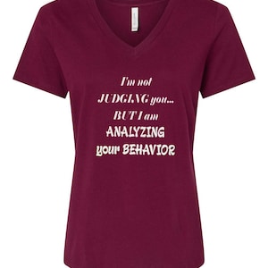 以下が含まれることがあります： 深紅色のVネックTシャツ。前面中央に白い文字で「I'm not JUDGING you... BUT I am ANALYZING your BEHAVIOR」と書かれています。Tシャツは半袖でシンプルなデザインです。