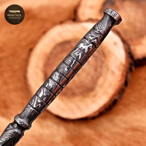 Puede incluir: Primer plano de un mango de cuchillo de acero de Damasco con un patr&oacute;n &uacute;nico. El mango es cil&iacute;ndrico con un agarre texturizado y un extremo acampanado. El logotipo "HERITAGE DAMASCUS" est&aacute; en la esquina superior izquierda. Fondo de madera.