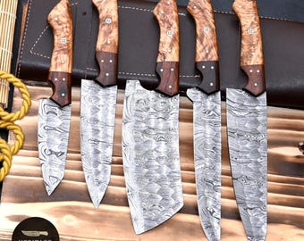 May include: A set of five Damascus steel knives with wooden handles. The blades have a unique patterned design. The handles are a light brown color with dark brown accents. The knives are displayed on a wooden surface.
